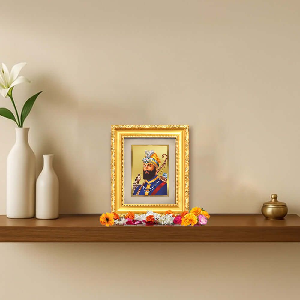 Guru Gobind Singh Ji framed portrait in 24K gold plated foil on a wooden shelf with flowers.